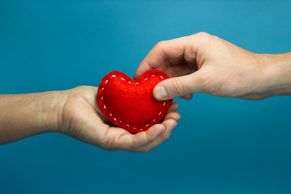 Caregiver Concept. Man Hand Puting The Red Soft Heart In Woman Hand On Blue Background